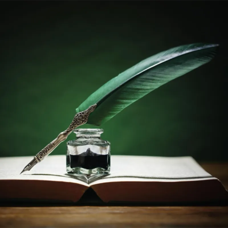 Grüne Schreibfeder und Tintenfass liegen auf einem aufgeschlagenen Buch vor dunkelgrünem Hintergrund.