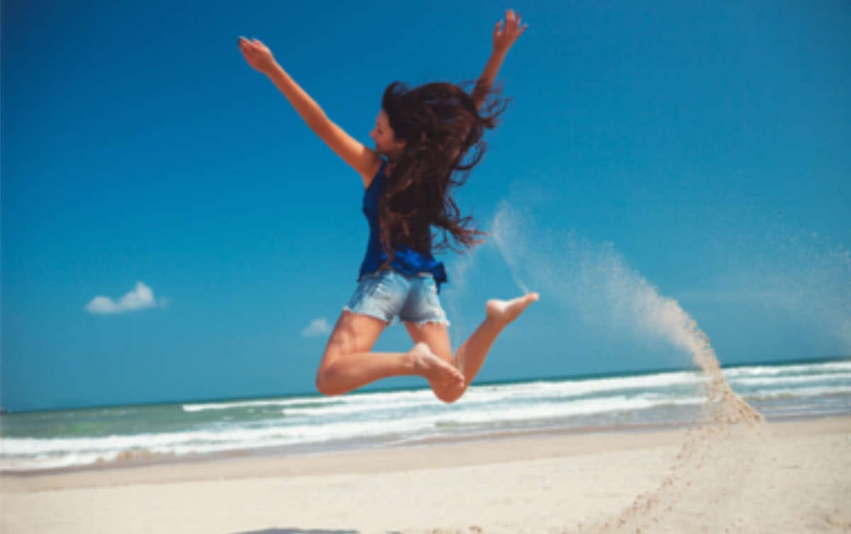 Innere Stärke Eine junge Frau in Jeans Shorts springt mit ausgebreteiteten Armen an einem Sandstrand in die Luft und freut sich des Lebens.