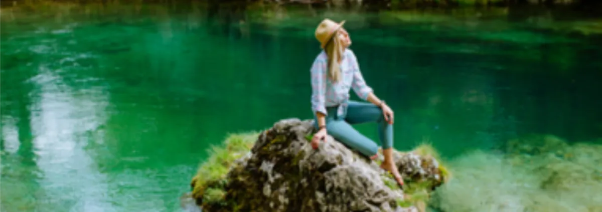 level Eine junge Frau sitzt auf einem mit Moos bewachsenen Stein in einem Weiher und schaut in den Himmel