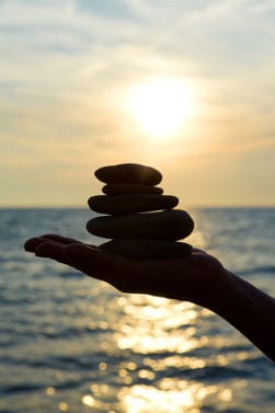 staerkefinden Eine Hand hält aufeinander aufgetürmte Kiesel gegen die Sonne, die über dem Meer untergeht