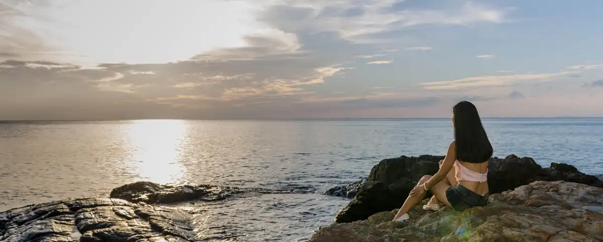 5-steps Frau blickt von einem Felsen am Ufer auf das Meer hinaus. Der Himmel ist wolkenverhangen, Dämmerung.