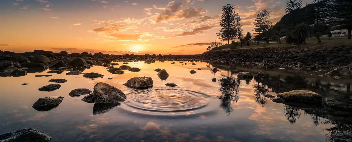 lake Steine mit sich ausbreitenden Wellen in einem See am rechten, abschüssigen Ufer unterhalb einer weideartigen Terasse. Sonneneuntergang im Hintergrund.
