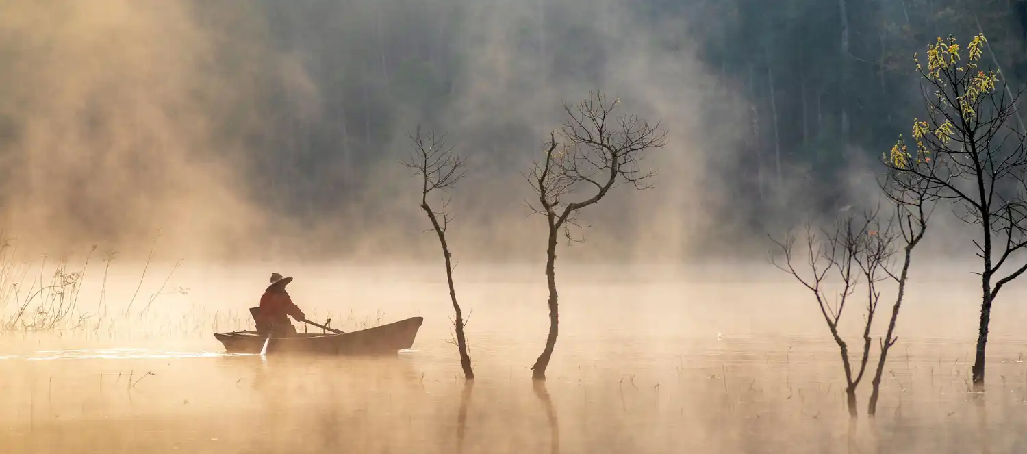 vertrauen Asiatischer Fischer im Ruderboot auf einem See, aus dem einige Laubfreie Bäume ragen. Nebel steigt über dem Wasser auf, diffuses Licht.