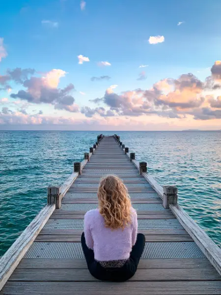 Eine Frau sitzt auf einem Holzsteg und blickt hinaus auf's Meer. Blauer Himmel mit einigen Wolken.
