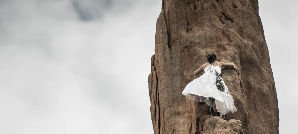 fehl-am-platz-wachstum Frau im Hochzeitskleid klettert auf einen Steilen Felsen. Betrachtung von hinten unten.