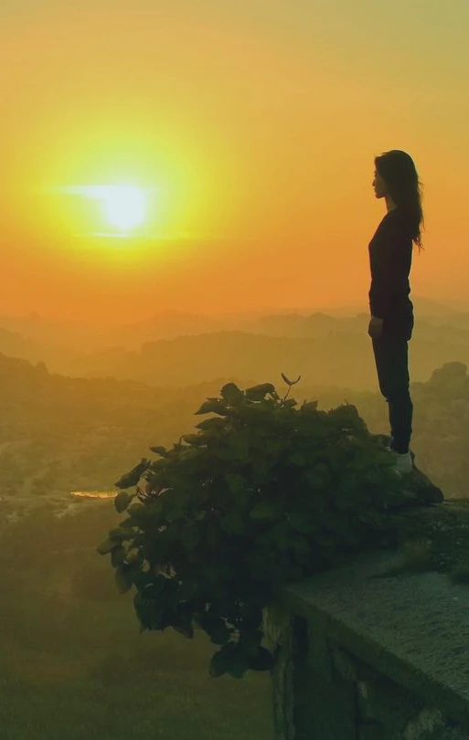 Eine Frau steht auf einer steinernen Brüstung im Sonnenuntergang und schaut über ein weites Tal