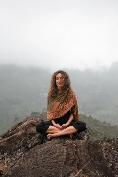 people-pleasing-frau-meditation Frau mit schwarzen Leggins und braun-orangem Umhang mit indischen Mustern sitzt im Schneidersitz auf einem Felsen und meditiert. Es ist neblig.