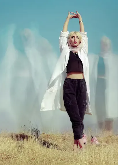 kinesiologie-sensibel Blonde Frau mit schwarzer Pludderhose, schwarzem Top und weissem Mantel streckt sich auf einer Wiese.