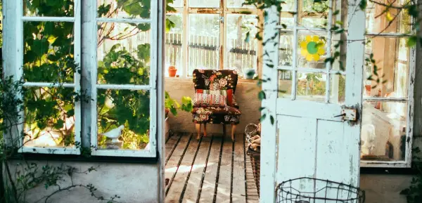 Blick durch die offene Tür eines gemütlichen, hellen Gartenhäuschens auf einen Patchwork-Sessel an der hinteren Wand unter der Fensterfront